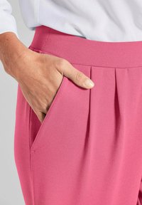 Roze, geplooide broeken met een gladde textuur en een zijzak, gecombineerd met een lichtgekleurde top die een deel van de mouw toont.