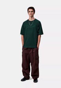 Ciemnozielona, oversizeowa koszulka z logo pisanym kursywą, zestawiona z luźnymi, brązowymi spodniami typu cargo z bocznymi kieszeniami i mankietem na dole nogawek.