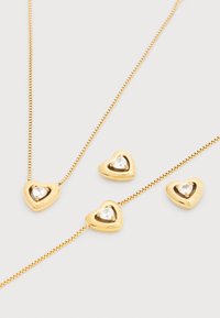 Κολιέ από επιχρυσωμένο μέταλλο με μενταγιόν σε σχήμα καρδιάς που διαθέτει ένα διαφανές πολύτιμο λίθο. Συνοδεύεται από τρία ταιριαστά μοτίφ καρδιάς, επίσης με πολύτιμους λίθους.