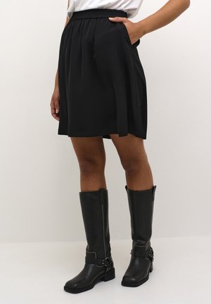 Woman wearing a black knee-length skirt with pockets and black knee-high boots with decorative straps and heels.