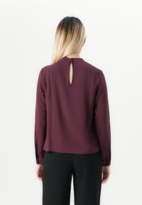 Kvinne med langt blondt hår iført en plommefarget langermet bluse med nøkkelhullsrygg og svarte bukser, stående mot en ensfarget hvit bakgrunn.