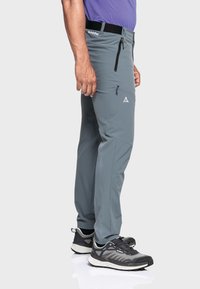 Grauweiche Softshell-Wandhose mit Reißverschlusstaschen, flexibel sitzend und aus atmungsaktivem Material, kombiniert mit schwarz-grauen Wanderschuhen.