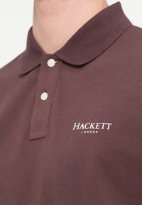 Tmavě hnědá polo tričko s žebrovaným límcem, se dvěma knoflíky a bílým logem "HACKETT LONDON" na levé horní straně.
