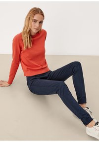 Pomarańczowy sweter o teksturowanej powierzchni z długimi rękawami, w zestawie z ciemnoniebieskimi, dopasowanymi spodniami. Białe sneakersy z czarnymi akcentami dopełniają strój.