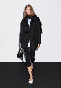 Donna che indossa un blazer nero e una sciarpa, una camicia azzurra chiara, leggings neri a 3/4, scarpe bianche e porta una piccola borsa nera contro un muro bianco.