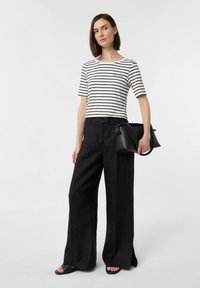 Schwarz-weiß gestreiftes T-Shirt mit kurzen Ärmeln, kombiniert mit lockeren schwarzen Hosen. Akzentuiert mit einem kleinen gestickten Logo und hält eine schwarze Tasche.