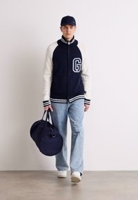 Námornícky a biely varsity kabát s písmenom "G", svetlomodré džínsy a námornícka športová taška. Model má na hlave námornícku čiapku a biele tenisky.