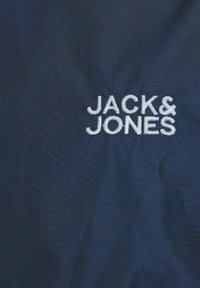 Mörkblått tyg med en strukturerad yta och en broderad vit logotyp "JACK & JONES" centrerad på materialet.