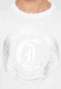 Hvit t-skjorte med et sølvfarget sirkulært slange design som omgir en stilisert "J" og "Just Cavalli"-tekst på midten av brystet.