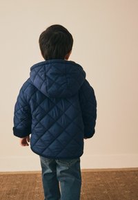 Quiltet navy blå jakke med hætte, prydet med diamantmønster og ribbede manchetter, parret med blå jeans mod en ensfarvet baggrund.