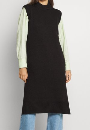 Vrouw draagt een mouwloze zwarte gebreide midijurk over een lichtgroen shirt met lange mouwen en lichtblauwe spijkerbroek, staande tegen een effen achtergrond.