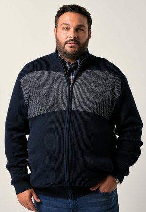 Marineblauer Reißverschluss-Pullover mit strukturiertem Strick und einem grauen gemusterten Einsatz. Verfügt über Seitentaschen und gerippte Bündchen für einen guten Sitz. Getragen über einem karrierten Hemd.