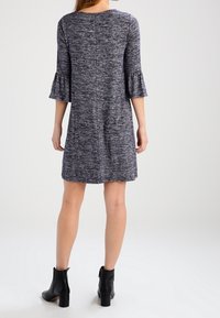 Graues, gestricktes Kleid mit strukturiertem Muster, drei Viertel-Glöckchenärmeln und geradem Schnitt. Getragen mit schwarzen Stiefeletten.