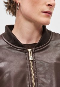 Brun læder bomberjakke med ribbet krave, har en guld lynlås og glat tekstur. Inkluderer et mørkebrunt strikket lag nedenunder.