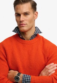 Suéter naranja brillante con textura acanalada, cuello redondo y cuello de camisa a cuadros azul asomando. Reloj negro con esfera plateada en la muñeca.