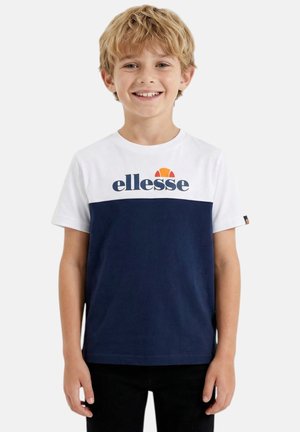 Garçon aux cheveux blonds souriant, portant un t-shirt à manches courtes blanc et bleu marine avec le logo « ellesse », debout devant un fond uni.