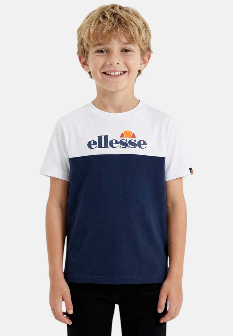 Garçon aux cheveux blonds souriant, portant un t-shirt à manches courtes blanc et bleu marine avec le logo « ellesse », debout devant un fond uni.