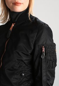 Veste bomber noire avec une finition lisse et brillante. Présente une fermeture éclair sur le devant, un col côtelé et une poche zippée sur la manche.