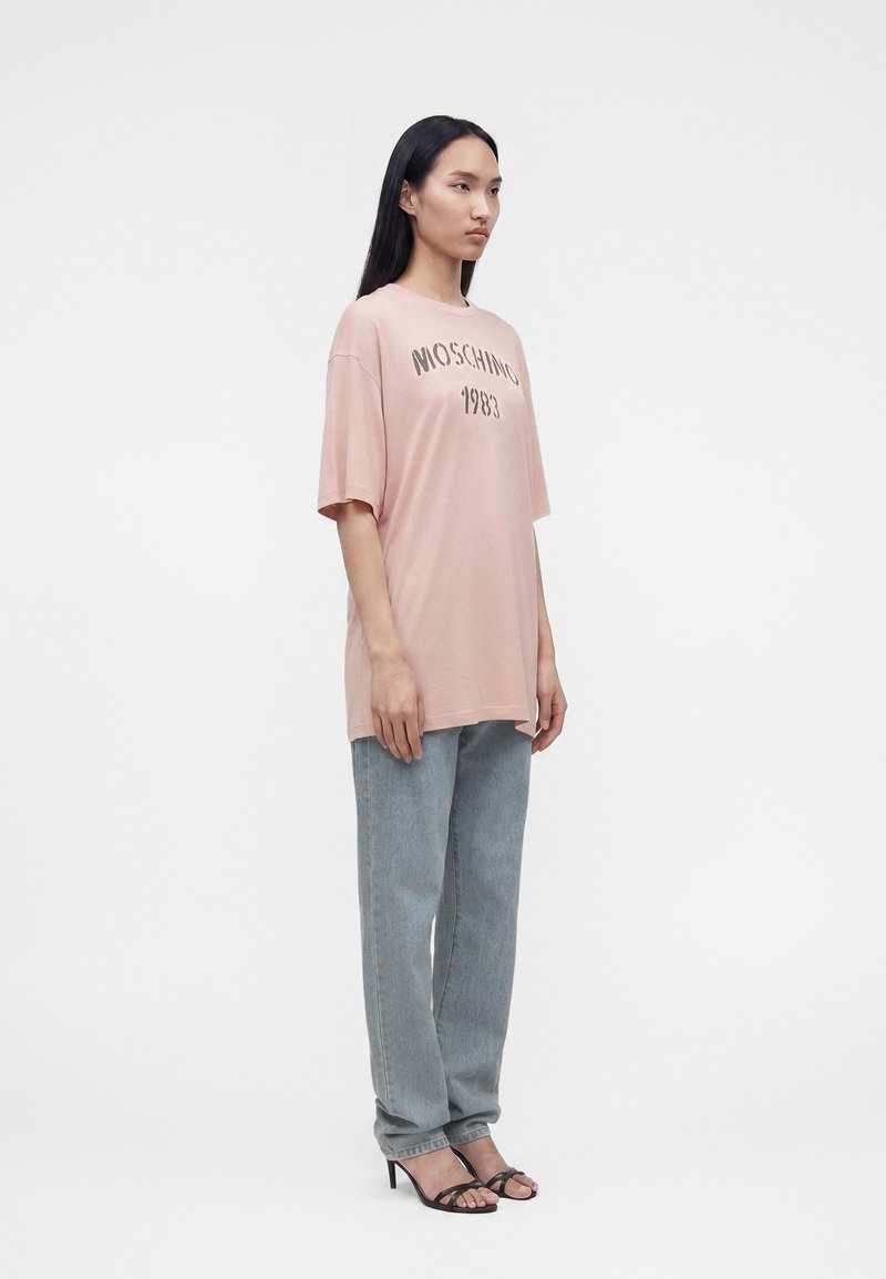 Woman with long black hair wearing oversized pink Moschino 1983 t-shirt, light blue straight-leg jeans, and black high-heeled sandals.