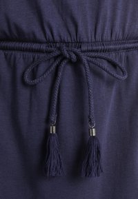 Tissu bleu marine avec une ceinture élastique nouée en nœud, comprenant deux cordons tressés se terminant par des glands avec embouts métalliques.