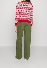Donna che indossa un maglione natalizio rosso e bianco con motivo di renne e pantaloni larghi verdi oliva, in piedi di spalle.
