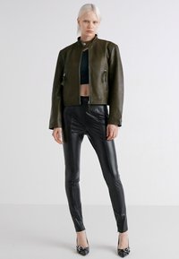 Giacca in pelle verde oliva con colletto alto, tasche con zip, abbinata a pantaloni in pelle neri aderenti e tacchi neri a punta.