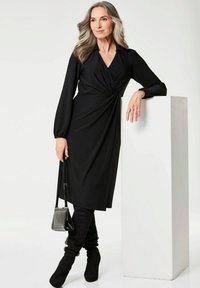 Schwarzes langärmliges Kleid mit einer Twist-Knoten-Detail an der Taille, kombiniert mit kniehohen schwarzen Wildlederstiefeln und einer kleinen schwarzen Handtasche.