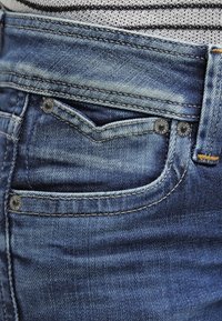 Jeans de mezclilla azul oscuro con zonas desgastadas, costuras visibles, un botón metálico y un bolsillo frontal con un acabado limpio.