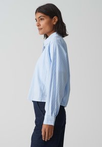 Camicia button-up a righe blu chiaro con colletto, design cropped e maniche lunghe, dotata di una texture di tessuto liscia e dettagli sui polsini.