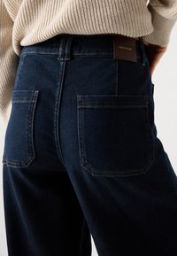 Pantalones de mezclilla de tiro alto en azul oscuro con bolsillos traseros; presentan costuras contrastantes y una etiqueta de cuero marrón en la cinturilla.