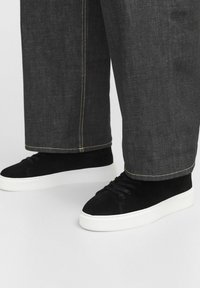 Baskets montantes en daim noir avec des semelles en caoutchouc blanches, dotées de lacets ronds et de surpiqûres contrastées visibles sur un pantalon en denim sombre.