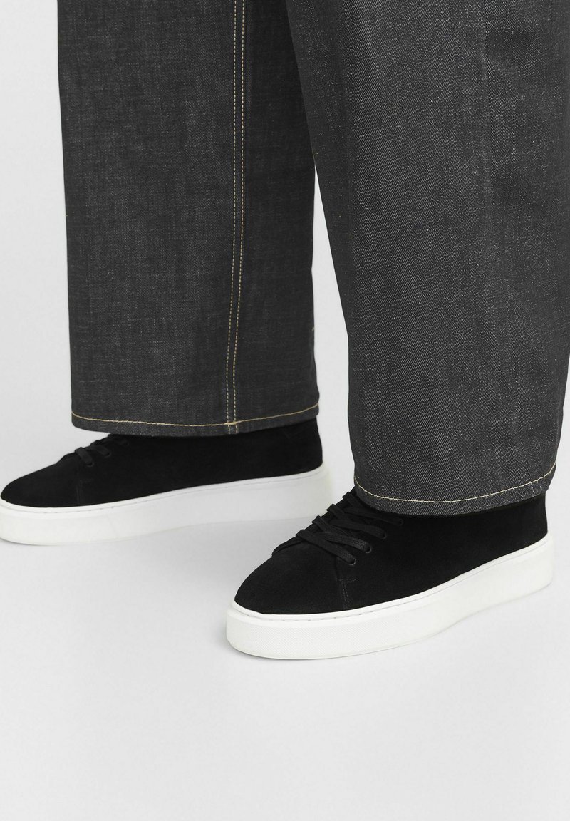 Baskets montantes en daim noir avec des semelles en caoutchouc blanches, dotées de lacets ronds et de surpiqûres contrastées visibles sur un pantalon en denim sombre.
