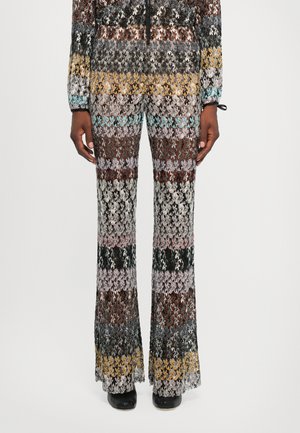 Person wearing multicolor crochet knit pants with a zigzag floral pattern and black heeled shoes, standing against a white background.