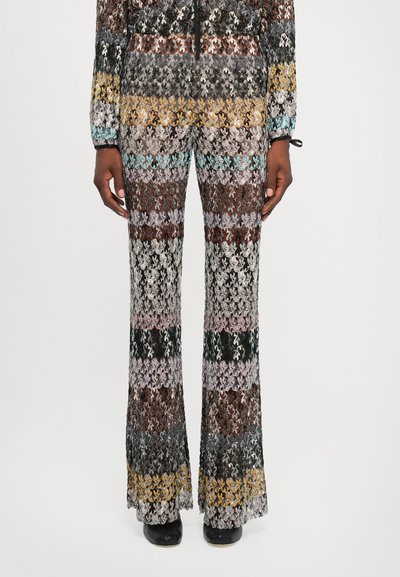 Person, die farbenfrohe gehäkelte Hosen mit einem Zickzack-Blumenmuster und schwarzen High Heels trägt, vor einem weißen Hintergrund steht.