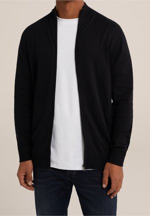Homme portant un cardigan noir zippé par-dessus un t-shirt blanc et un jean bleu foncé, debout devant un fond neutre.