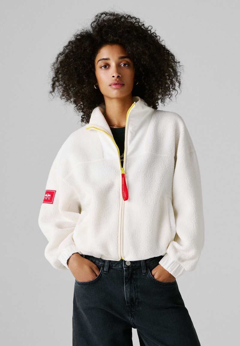 Λευκό φλις μπουφάν με ψηλό γιακά, κίτρινο φερμουάρ, κόκκινη λεπτομέρεια στο μανίκι και ελαστικά τελειώματα στα μανίκια· συνδυασμένο με μαύρα τζιν.