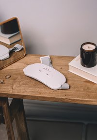 Stylpro-Gerät aus weißem Kunststoff mit grauem Band, ausgestattet mit Tasten und Branding. In der Nähe befinden sich eine Kerze und ein Stapel Bücher auf einem Holztisch.