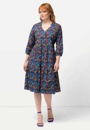 Blumenkleid mit Marineblauem Hintergrund, V-Ausschnitt, dreiviertel Ärmeln und gestuftem Rock. Getragen mit blauen Riemchen-Heels.