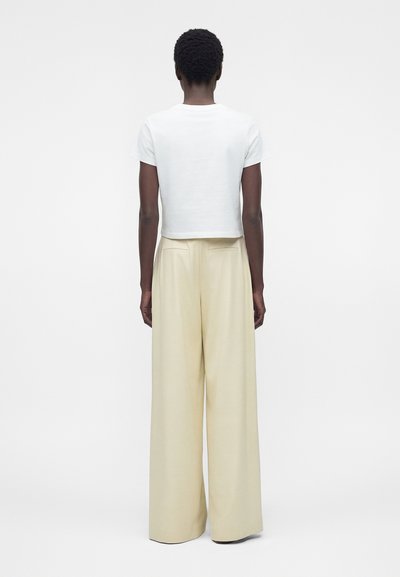 Άτομο με κοντά κατσαρά μαλλιά στέκεται με την πλάτη στραμμένη, φοράει λευκό μπλουζάκι με κοντά μανίκια και εκρού παντελόνια με φαρδιά πόδια.