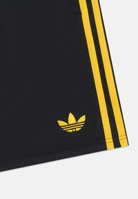 Calças curtas atléticas pretas com um design de três riscas amarelas nas laterais. Apresenta um logo bordado amarelo no canto inferior esquerdo. Textura suave.