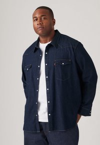 Cămașă din denim albastru închis cu un design western clasic, având umeri ascuțiți, nasturi tip snap și două buzunare pe piept. Mâneci lungi, croială lejeră.