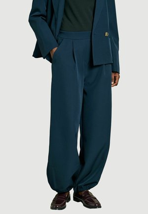 Person trägt dunkelblaue, lockere Hose mit elastischen Bündchen, passenden Blazer, schwarzes Hemd und dunkelbraune Lederloafers.