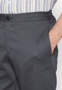 Šedé kalhoty na míru s plochým předním dílem, bočními kapsami a elastickým pasem se zapínáním na knoflík. Hladká textilie.