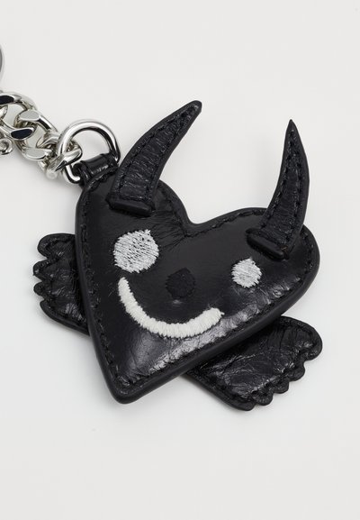 Clesă din piele neagră în formă de inimă, cu accesorii din metal argintiu, având ochi și zâmbet albe brodate, precum și mici coarne și aripi.