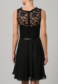 Robe noire avec un haut en dentelle, sans manches, et une jupe évasée. Dispose d'une ceinture en satin à la taille et d'un motif en dentelle détaillé.