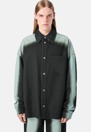 Joven con el cabello largo, vistiendo una camisa de botones negra y verde desgastada de corte oversized y pantalones a juego, contra un fondo liso.