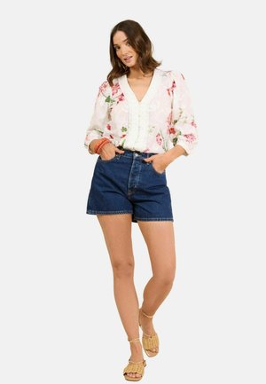 Nő virágmintás fehér blúzban, puffos ujjakkal, sötét farmerrövidnadrágban, fonott szandálban és narancssárga karkötőkkel, zsebekbe tett kézzel pózol.