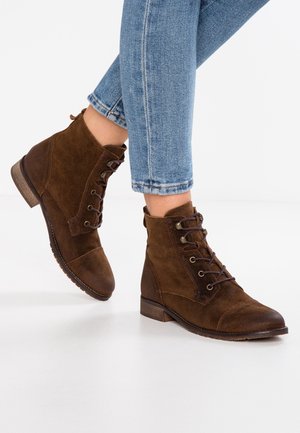 Bottines en cuir suédé marron, à bout rond, avec des semelles en caoutchouc et des œillets en métal. Elles sont dotées de lacets et d'un léger talon ; des coutures décoratives accentuent le design.
