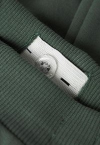 Tissu vert avec une texture côtelée, présentant une étiquette blanche avec un bouton et des coutures noires. Accent sur la zone du col.
