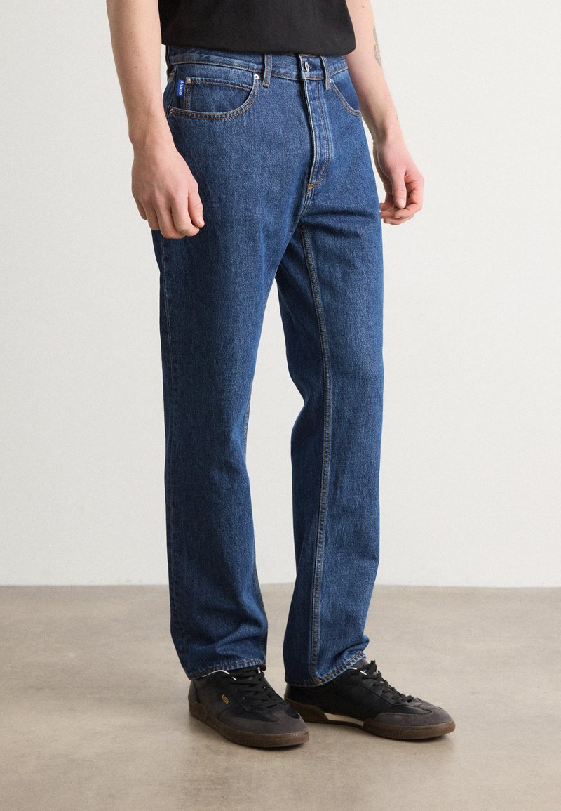 Jeans de mezclilla azul oscuro con corte recto, que presentan un diseño clásico de cinco bolsillos y costuras visibles, combinados con zapatillas negras.